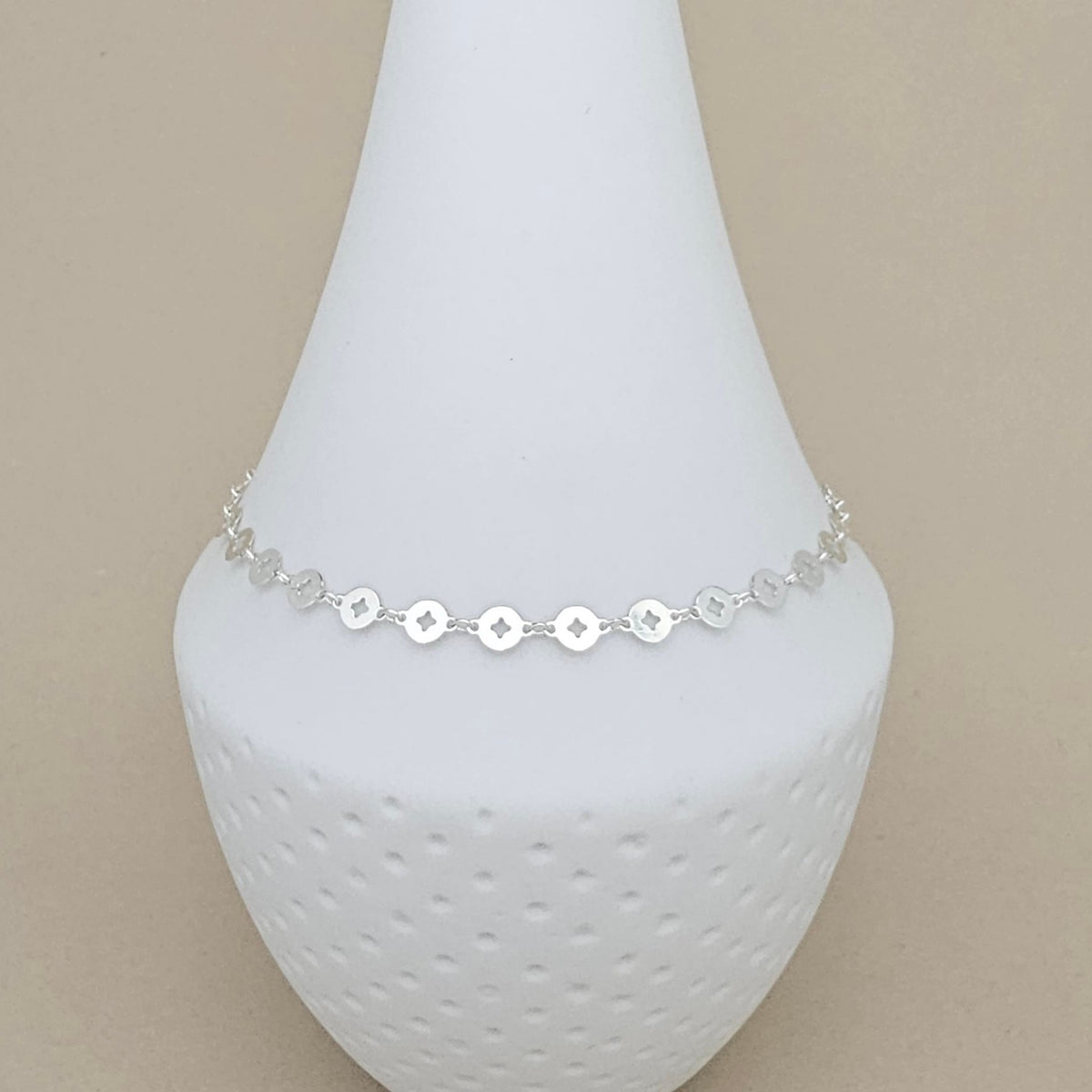 A delicate silver anklet with small circular disc charms and star details, displayed on a white textured cone stand against a solid beige background.