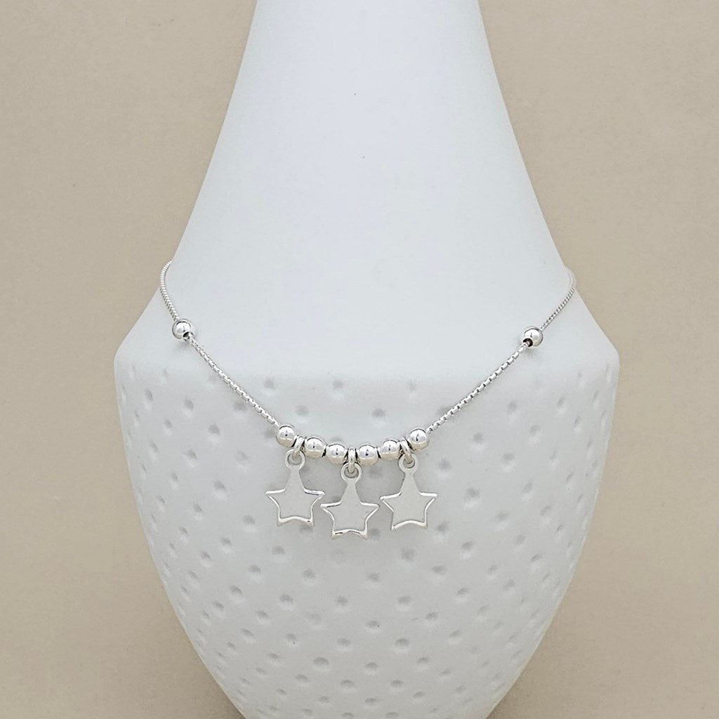 A silver anklet with three dangling star charms and small bead accents, displayed on a white textured jewelry bust against a beige background.
