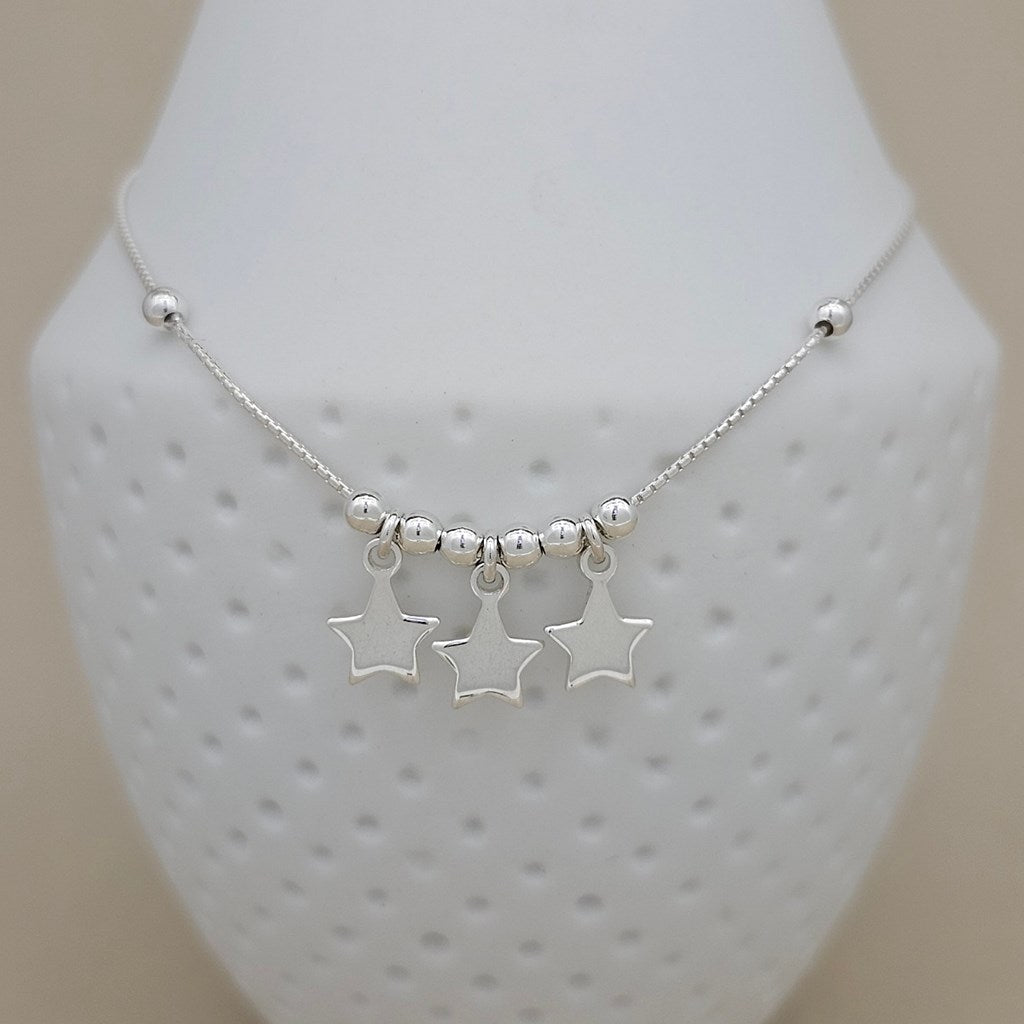 A silver anklet with three dangling star charms and small silver beads, displayed on a white textured jewelry bust against a light beige background.