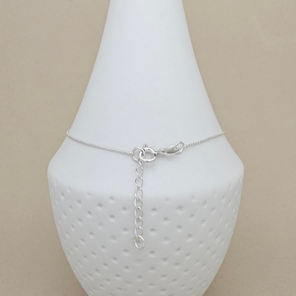 A delicate silver chain anklet with a lobster clasp and adjustable chain extension, displayed on a white textured cone jewelry stand against a beige background.
