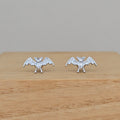 A pair of silver bat stud earrings with wings spread, displayed on a light wooden block against a neutral gray background.