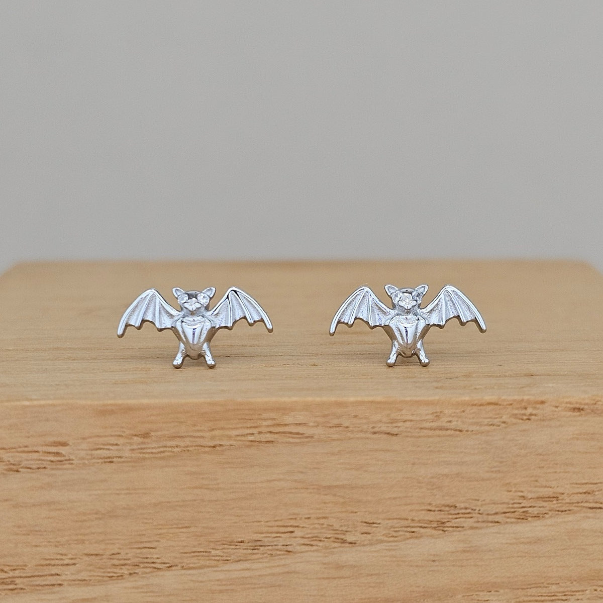 A pair of silver bat stud earrings with wings spread, displayed on a light wooden block against a neutral gray background.