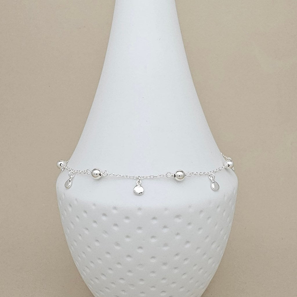 A delicate silver anklet with small bead and teardrop charms, displayed on a white textured cone stand against a beige background.