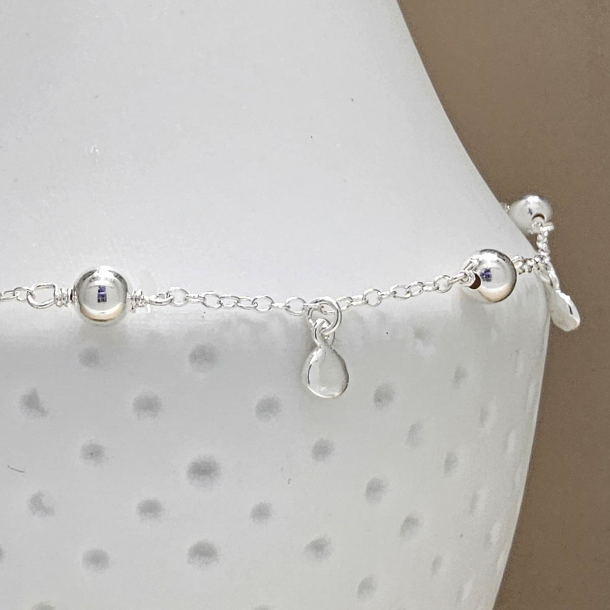 A delicate silver chain anklet with small polished beads and a teardrop charm, displayed on a white textured jewelry bust against a neutral beige background.