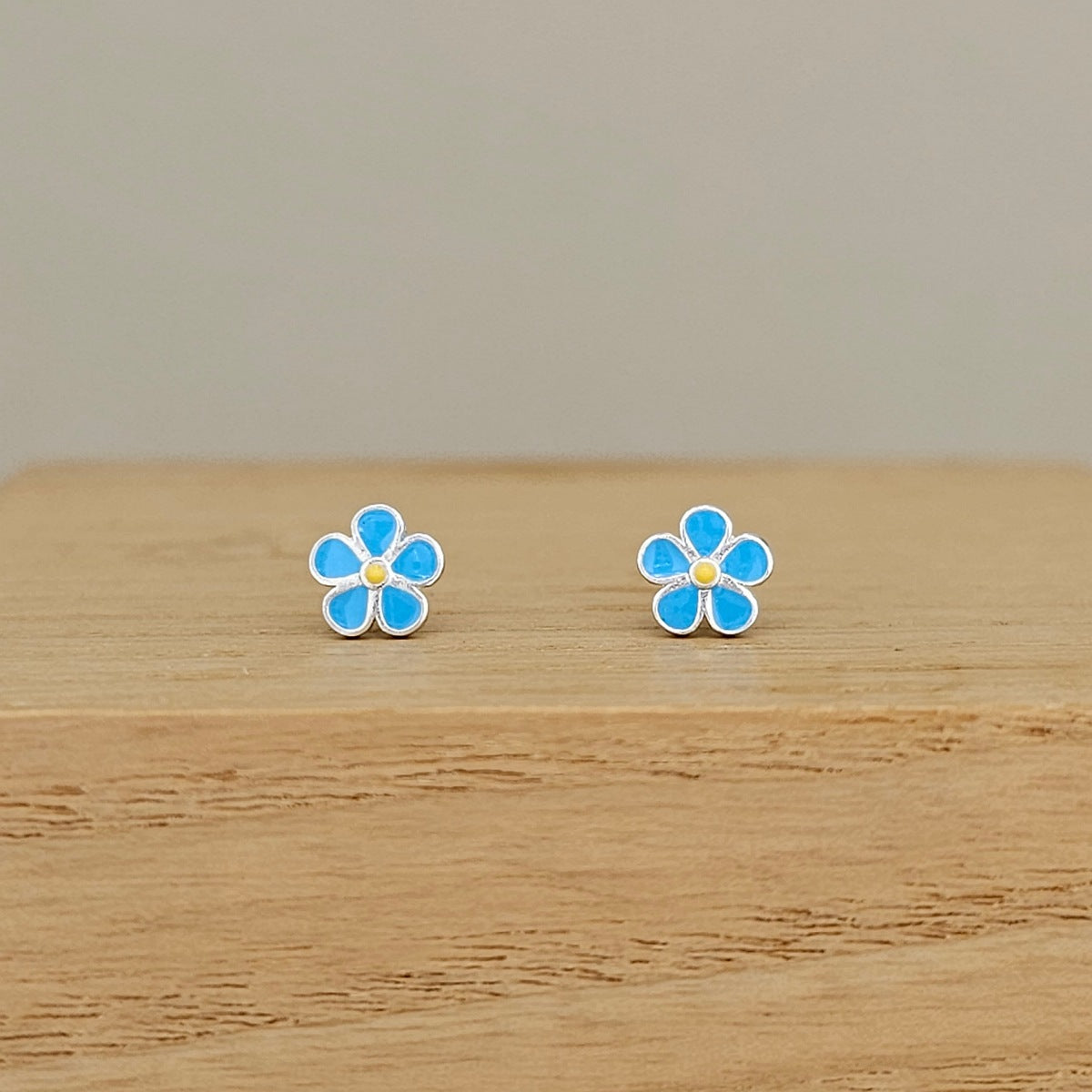 A pair of small flower-shaped stud earrings with bright blue enamel petals and gold-toned centers, displayed on a light wood surface against a soft beige background.
