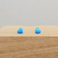 A pair of small, glossy light blue heart-shaped stud earrings displayed on a light wood block against a neutral gray background.