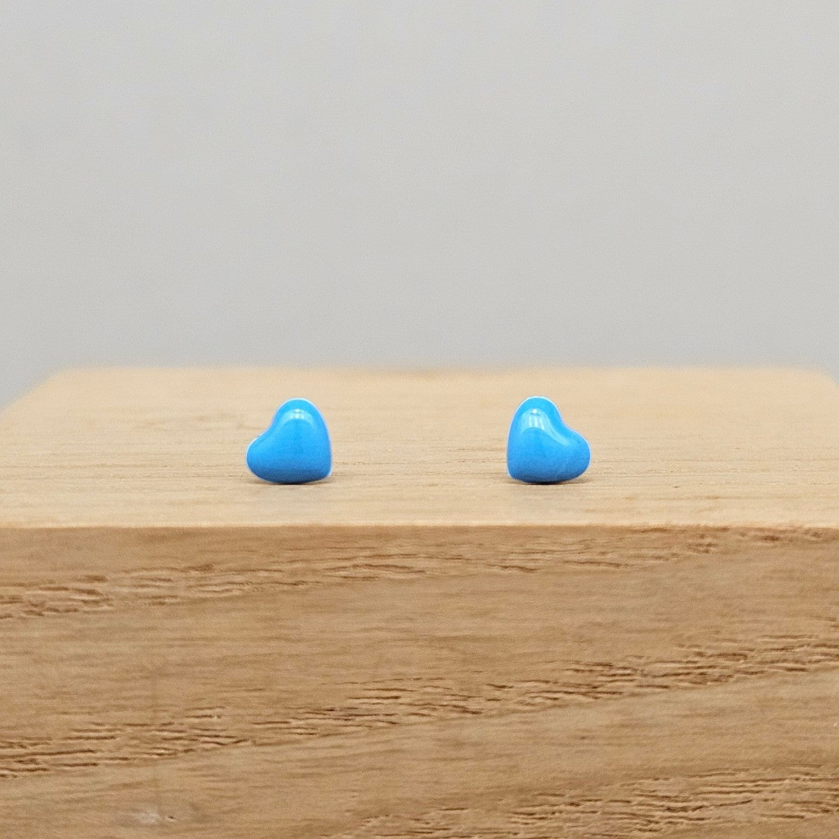 A pair of small, glossy light blue heart-shaped stud earrings displayed on a light wood block against a neutral gray background.