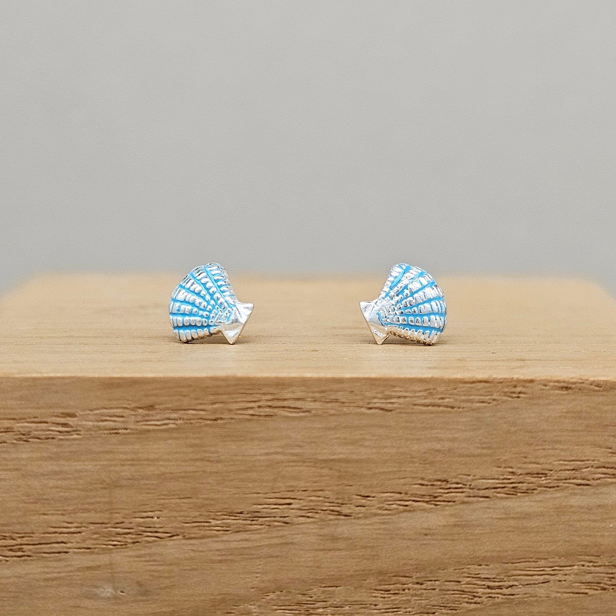 A pair of small shell-shaped stud earrings in light blue and white enamel with silver-toned posts, displayed on a wooden block against a plain gray background.