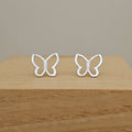 A pair of silver butterfly-shaped stud earrings with small clear stones in the center, displayed on a light wood block against a neutral beige background.