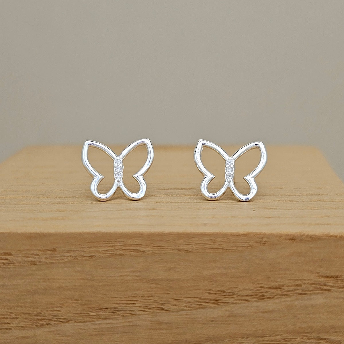 A pair of silver butterfly-shaped stud earrings with small clear stones in the center, displayed on a light wood block against a neutral beige background.