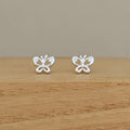 A pair of silver butterfly stud earrings with small clear stones, displayed on a wooden surface against a neutral beige background.
