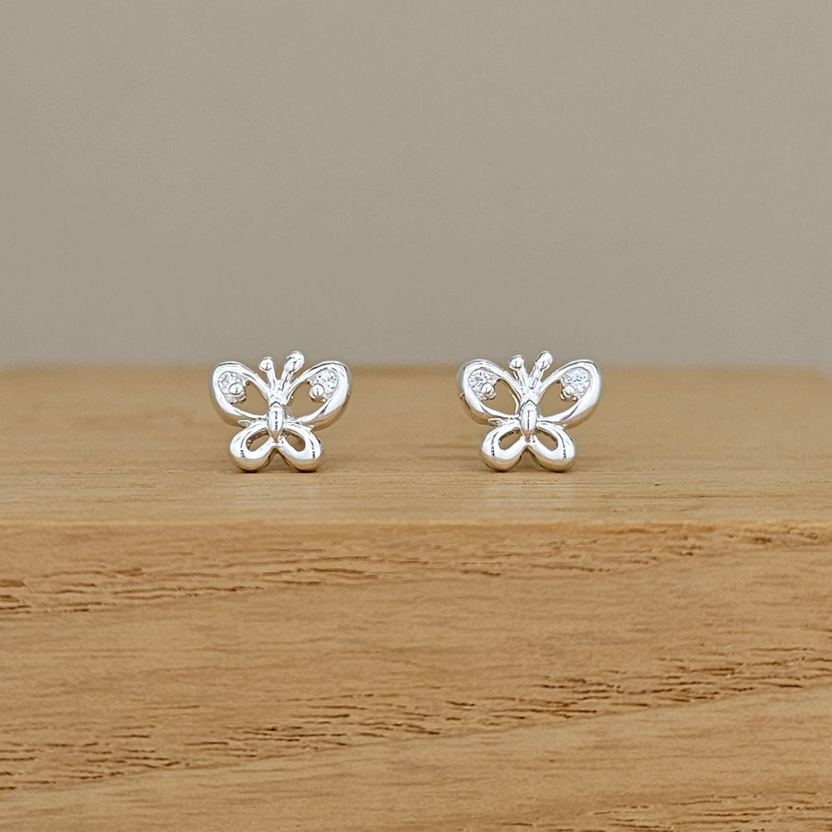 A pair of silver butterfly stud earrings with small clear stones, displayed on a wooden surface against a neutral beige background.
