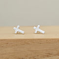A pair of silver cross stud earrings with a clear crystal in the center, resting on a light wooden surface against a neutral beige background.