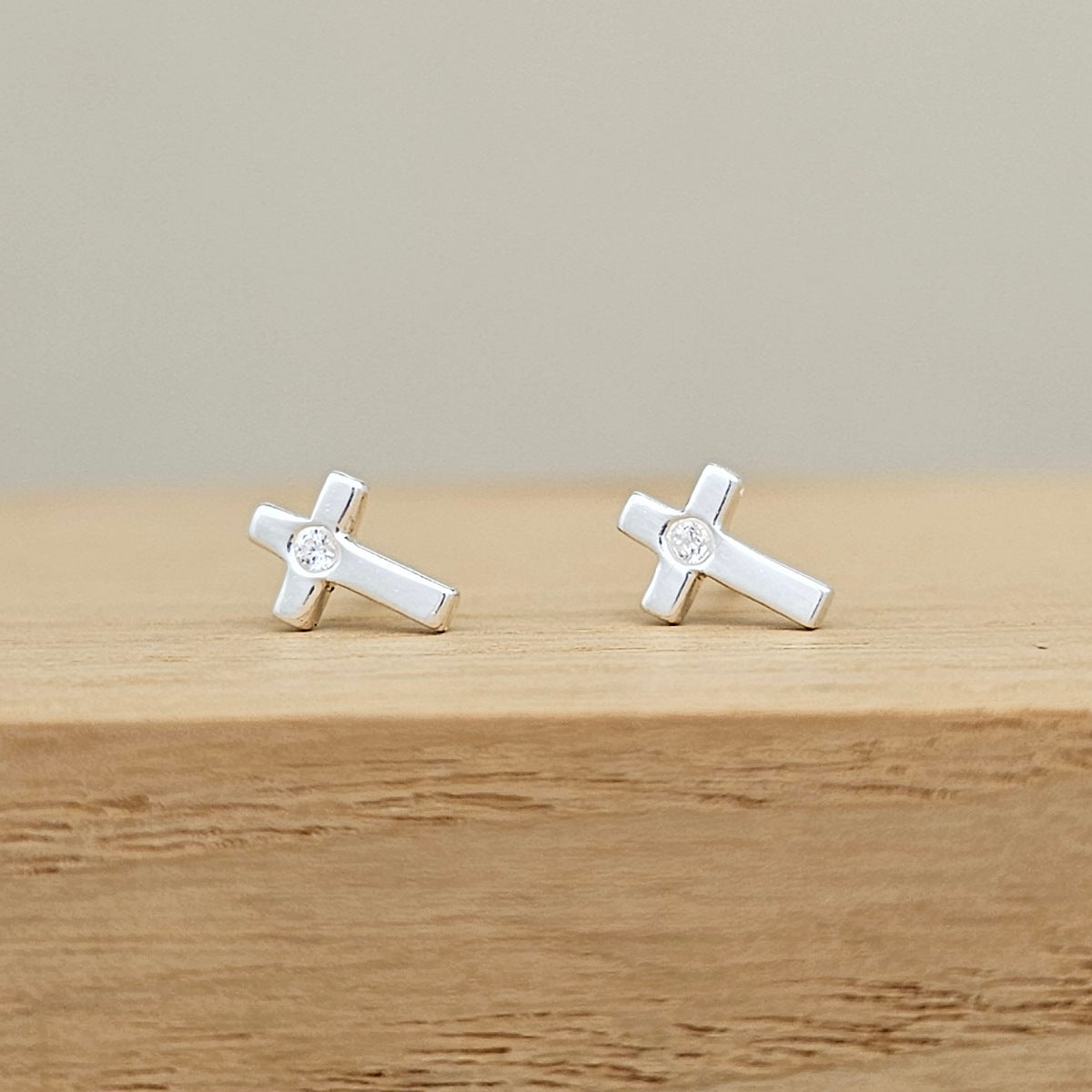 A pair of silver cross stud earrings with a clear crystal in the center, resting on a light wooden surface against a neutral beige background.