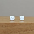 A pair of silver cat-face stud earrings resting on a light brown wooden surface against a neutral gray background.