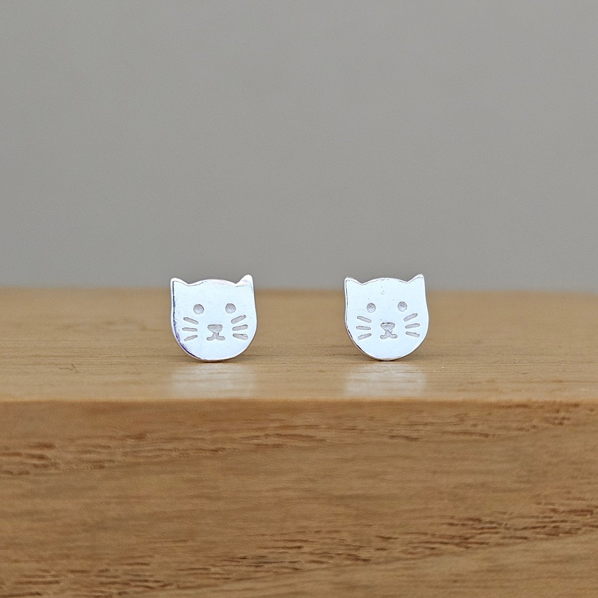 A pair of silver cat-face stud earrings resting on a light brown wooden surface against a neutral gray background.