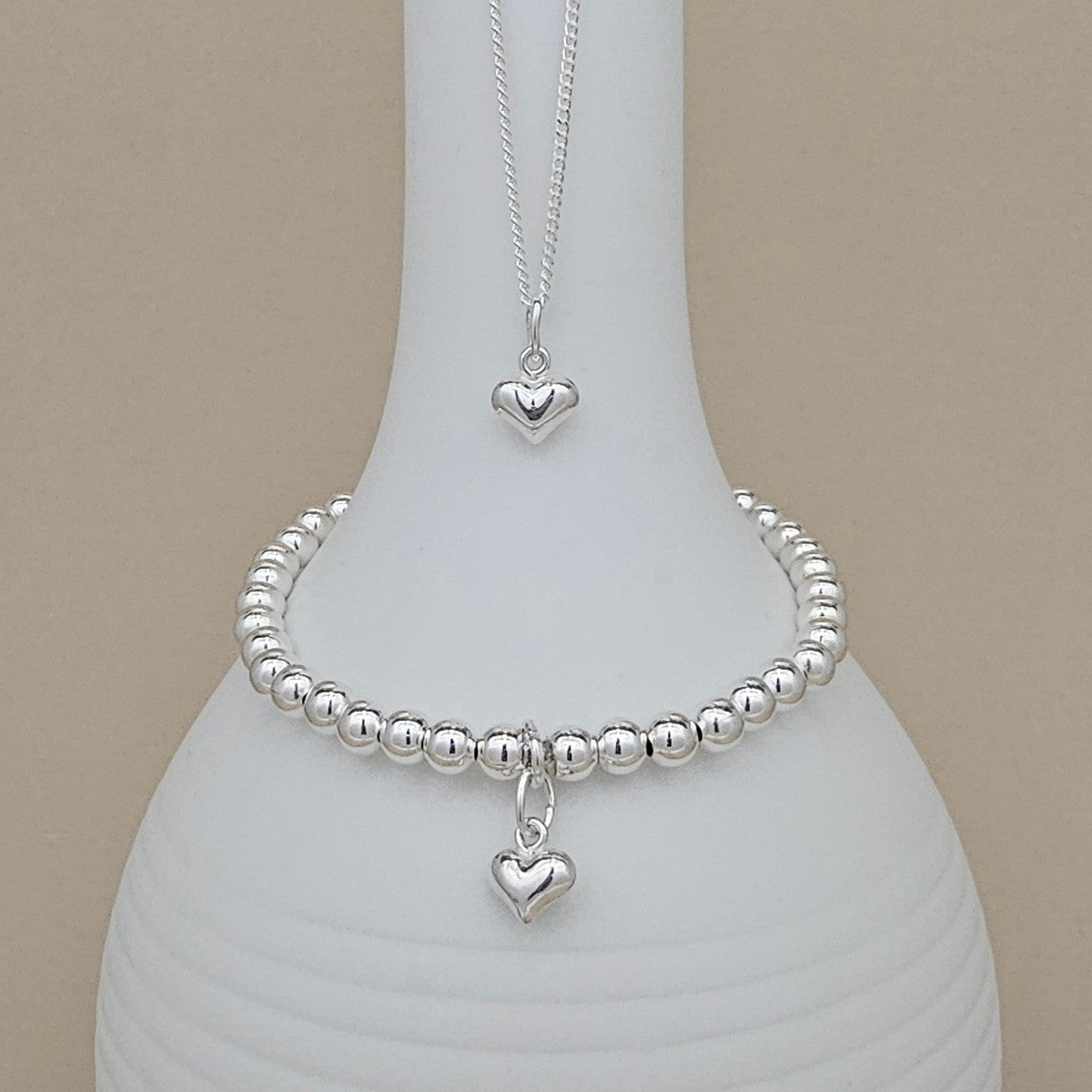 A silver beaded bracelet and matching necklace with heart charms, displayed on a white ceramic vase against a beige background.