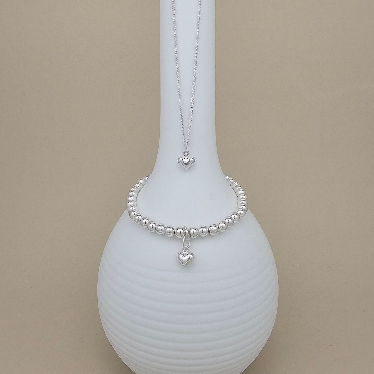 A silver beaded bracelet with a dangling heart charm is displayed on a white ribbed vase, with a matching silver heart pendant necklace hanging from the top of the vase, against a solid beige background.