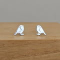 A pair of silver bird stud earrings with white enamel detailing, displayed on a wooden block against a neutral gray background.