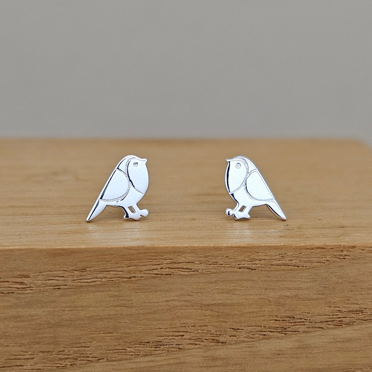 A pair of silver bird stud earrings with white enamel detailing, displayed on a wooden block against a neutral gray background.