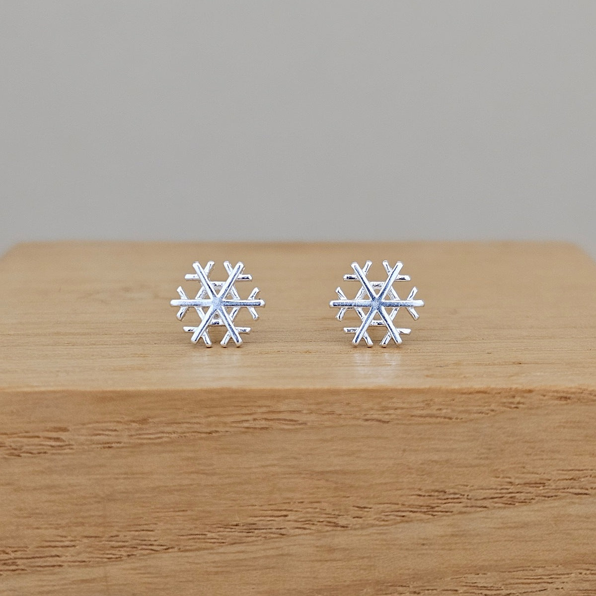 A pair of silver snowflake stud earrings resting on a light brown wooden block against a neutral gray background.
