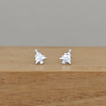 A pair of small silver stud earrings shaped like stylized trees, resting on a wooden surface against a plain gray background.