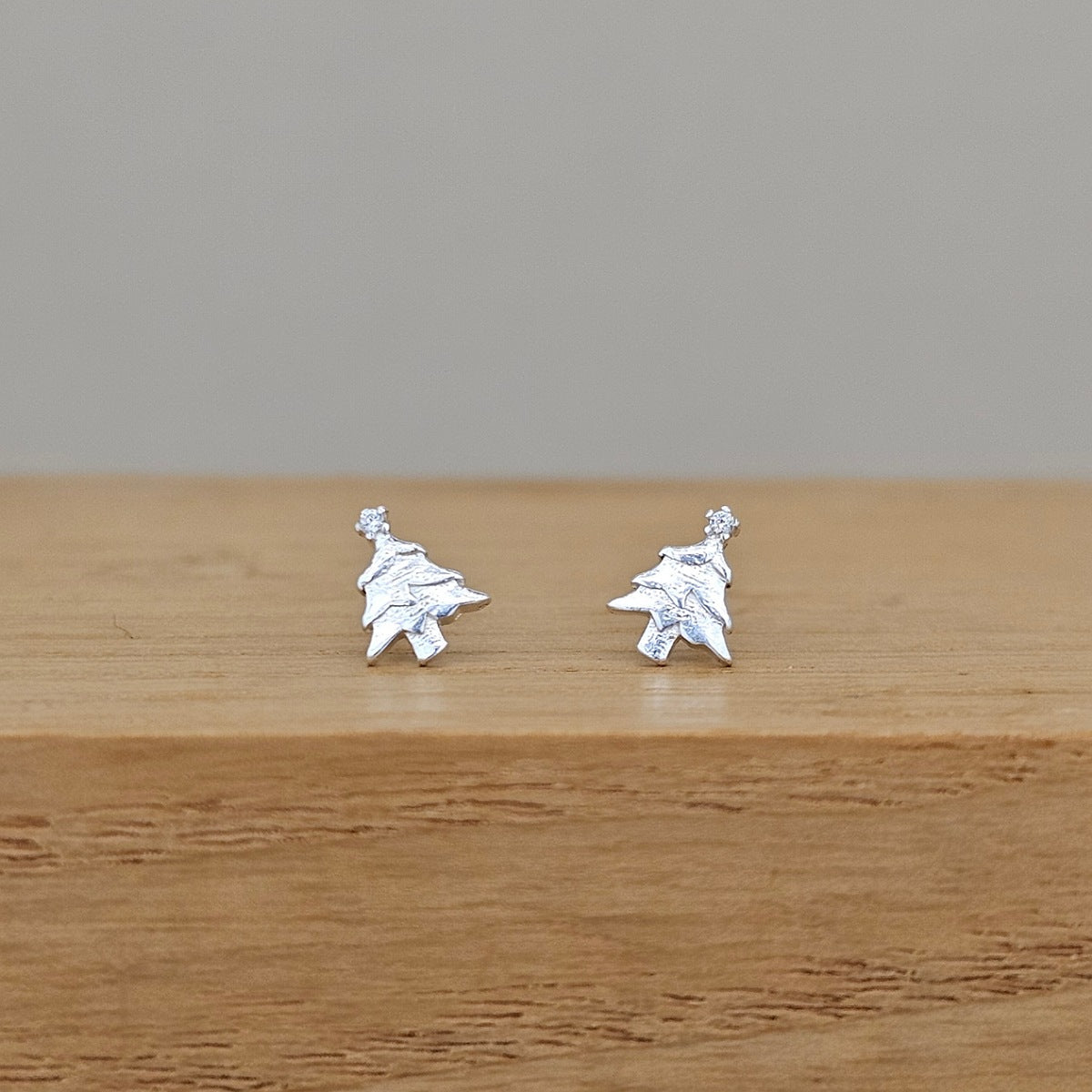 A pair of small silver stud earrings shaped like stylized trees, resting on a wooden surface against a plain gray background.