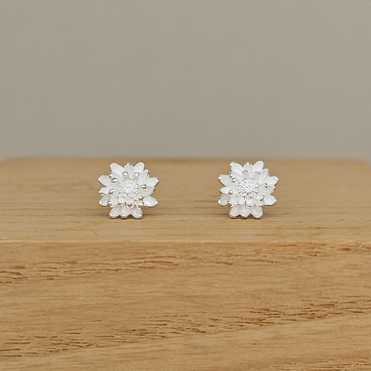 A pair of silver flower-shaped stud earrings displayed on a light wood surface against a neutral beige background.