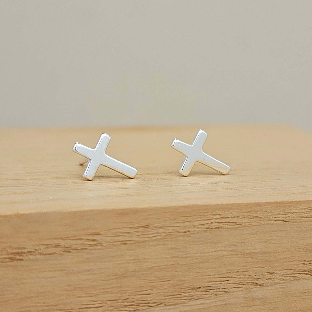 A pair of small silver cross stud earrings resting on a light wood surface against a soft beige background.