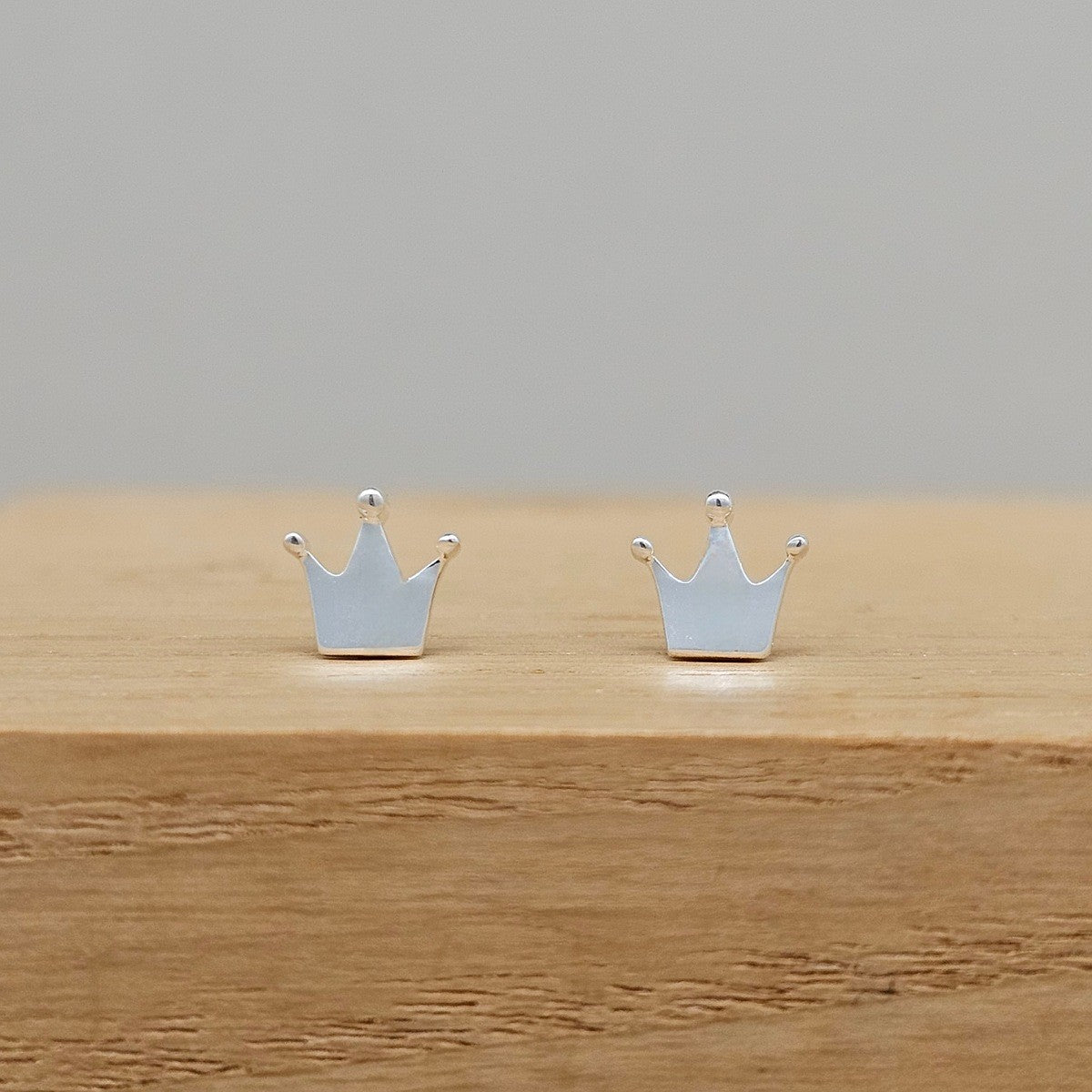 A pair of small silver crown stud earrings displayed on a light wood block against a neutral gray background.