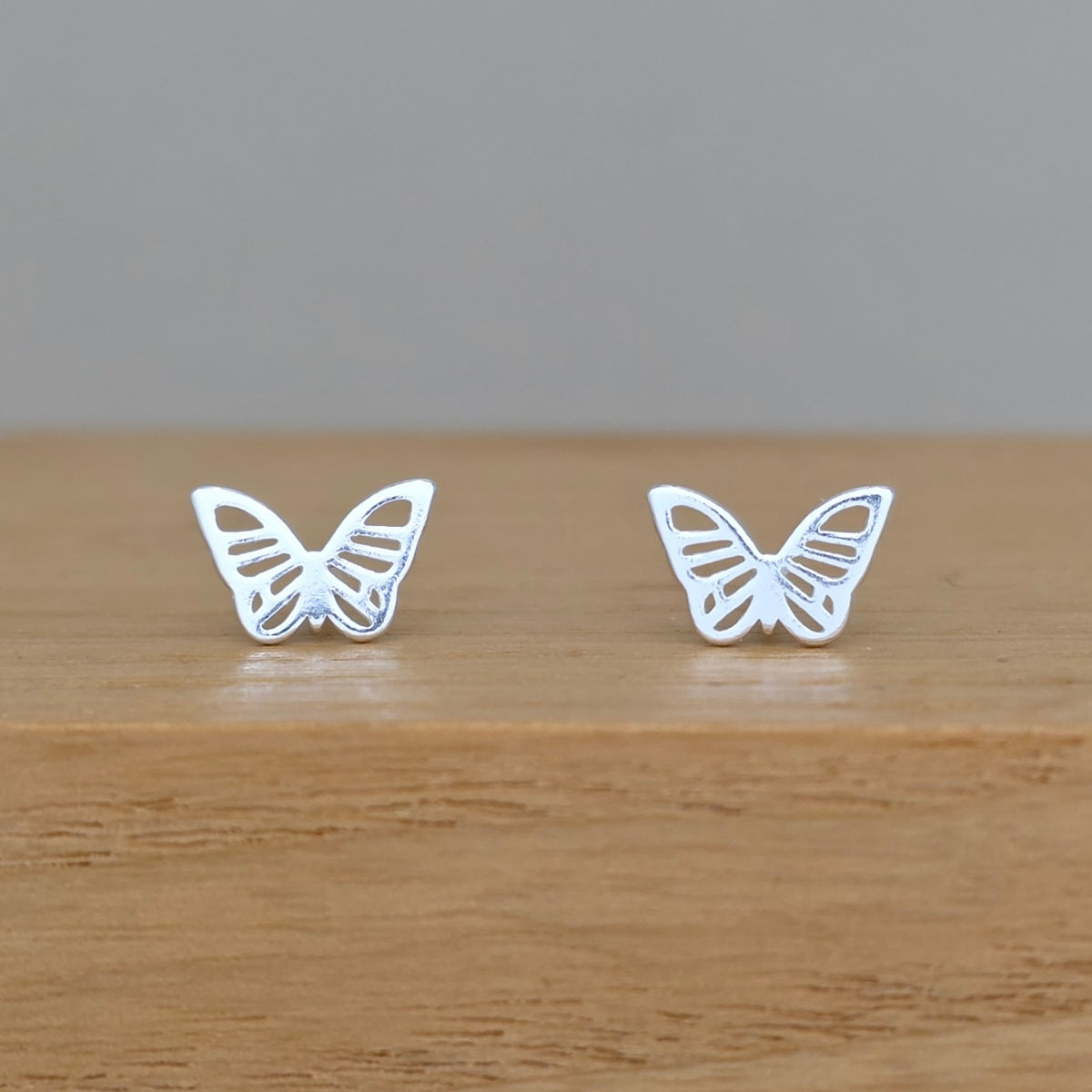 A pair of silver butterfly stud earrings displayed on a light brown wooden surface against a neutral gray background.