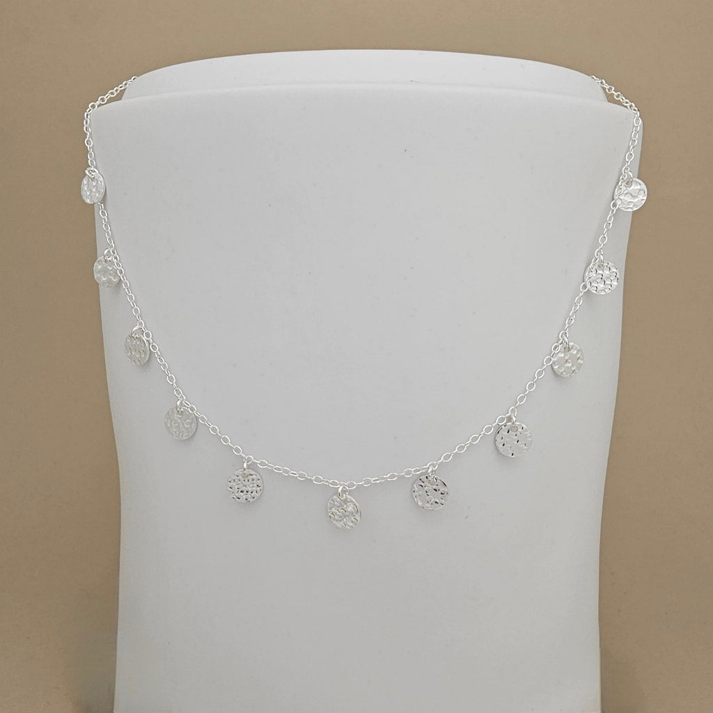 A delicate silver chain necklace with multiple small, circular, textured disc charms draped over a white display bust against a solid beige background.