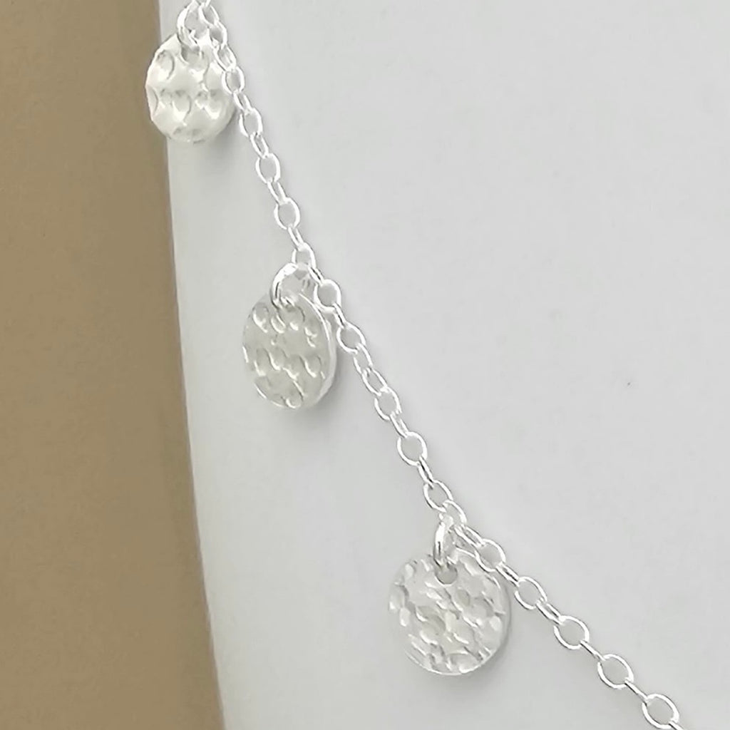 A close up of the silver chain necklace with three textured, hammered round disc charms, displayed on a neutral light gray background.