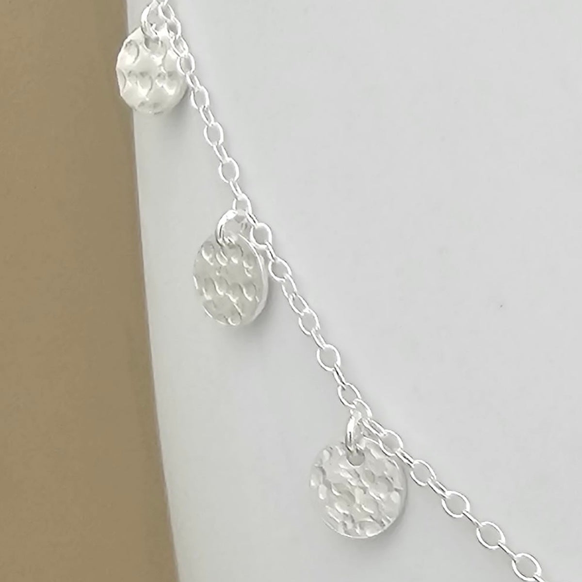 A close up of the silver chain necklace with three textured, hammered round disc charms, displayed on a neutral light gray background.