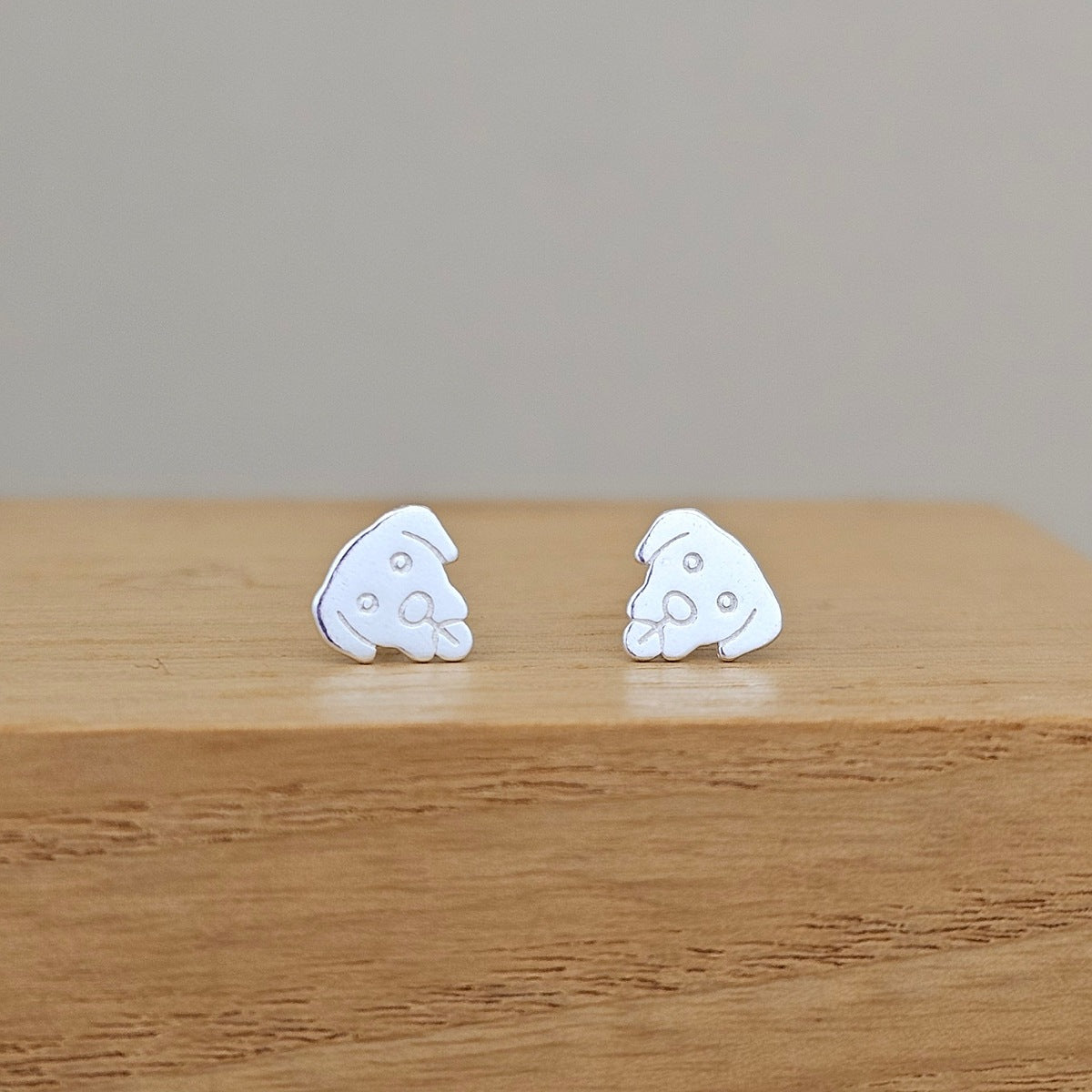 A pair of small white enamel stud earrings shaped like cartoonish dog faces, resting on a light brown wooden surface against a neutral gray background.