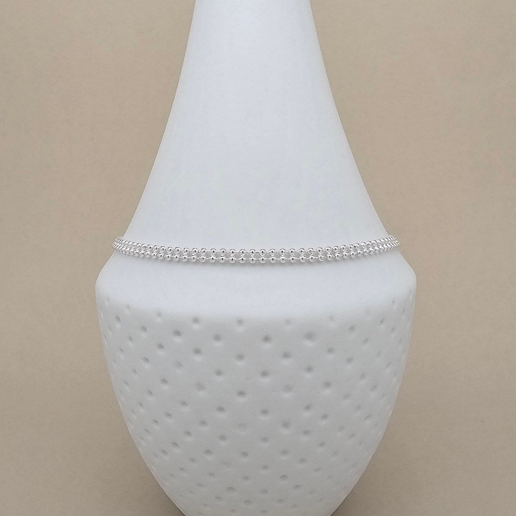 A delicate silver beaded anklet displayed on a white textured glass vase against a solid beige background.