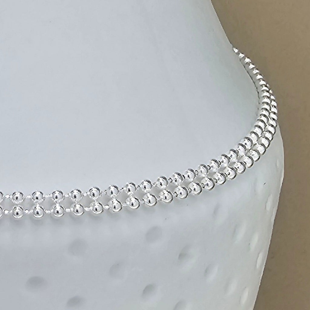 A close-up of a delicate silver beaded anklet draped over a white textured jewelry display bust.