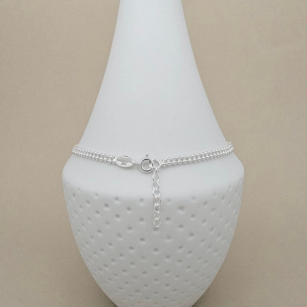 A delicate silver chain anklet with a small bead section, a clear faceted bead accent, and an adjustable lobster clasp, displayed on a white textured jewelry bust against a beige background.