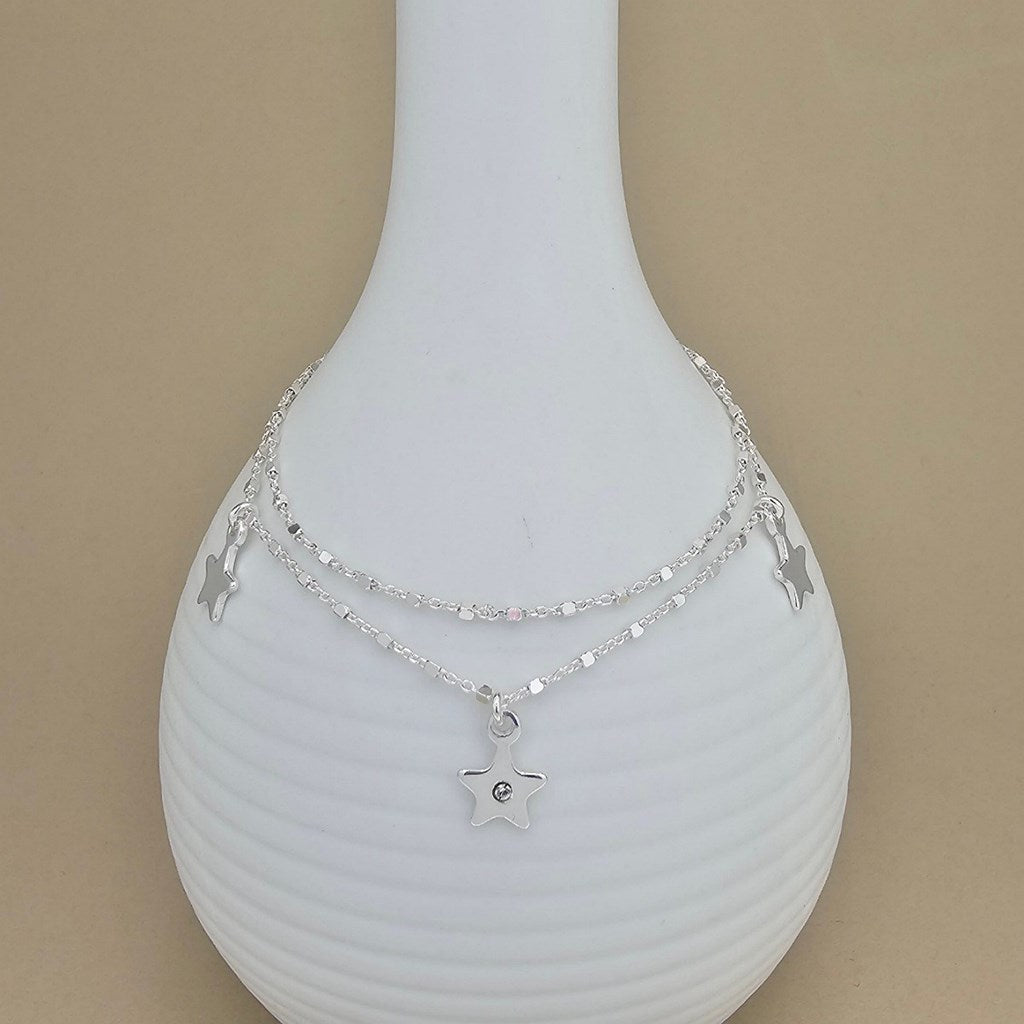 A delicate silver double-strand bracelet with star charms, displayed on a ribbed white ceramic vase against a solid beige background.