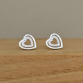 A pair of silver heart stud earrings with a double-heart design, one featuring a small pave-set heart in the center, displayed on a wooden block against a neutral background.