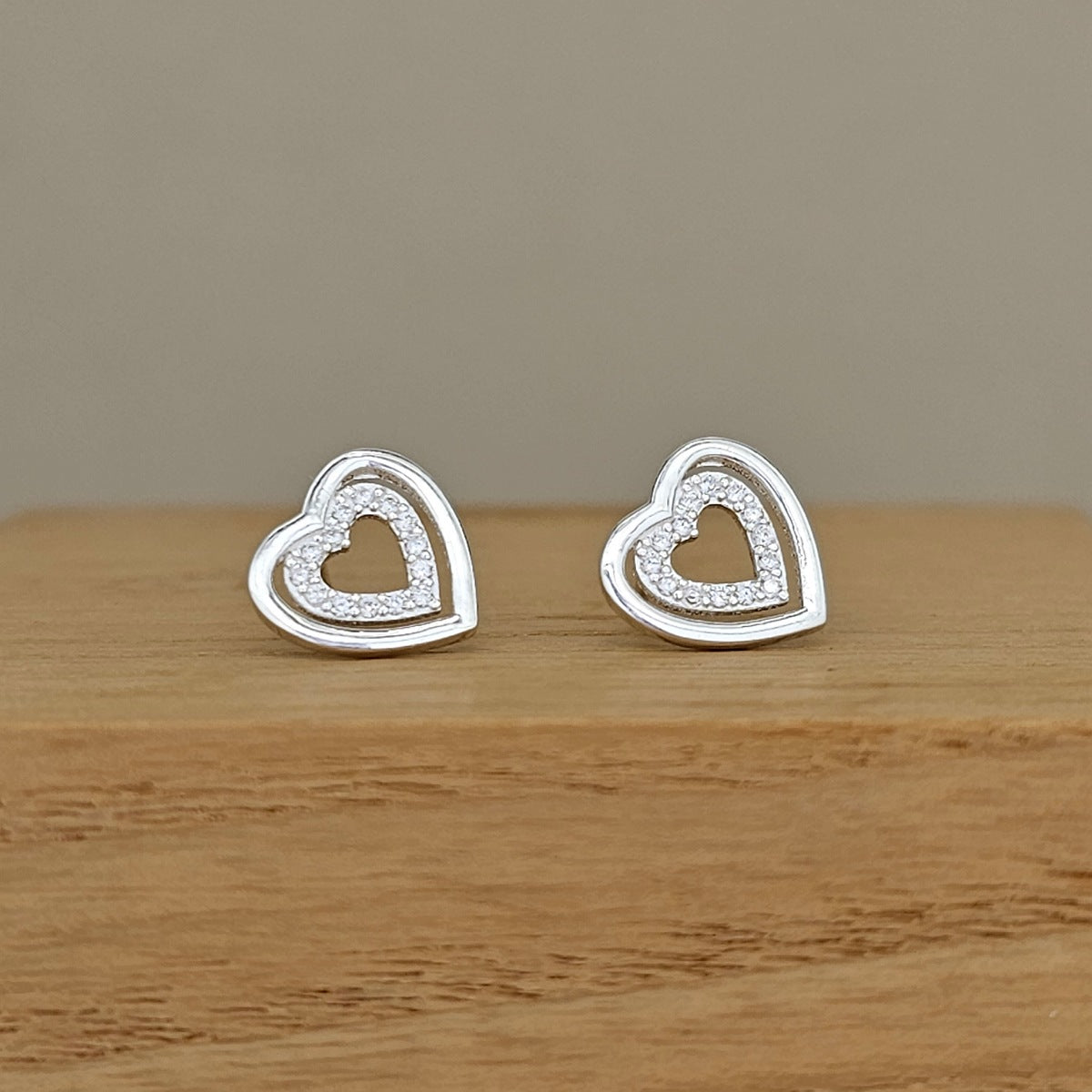 A pair of silver heart stud earrings with a double-heart design, one featuring a small pave-set heart in the center, displayed on a wooden block against a neutral background.