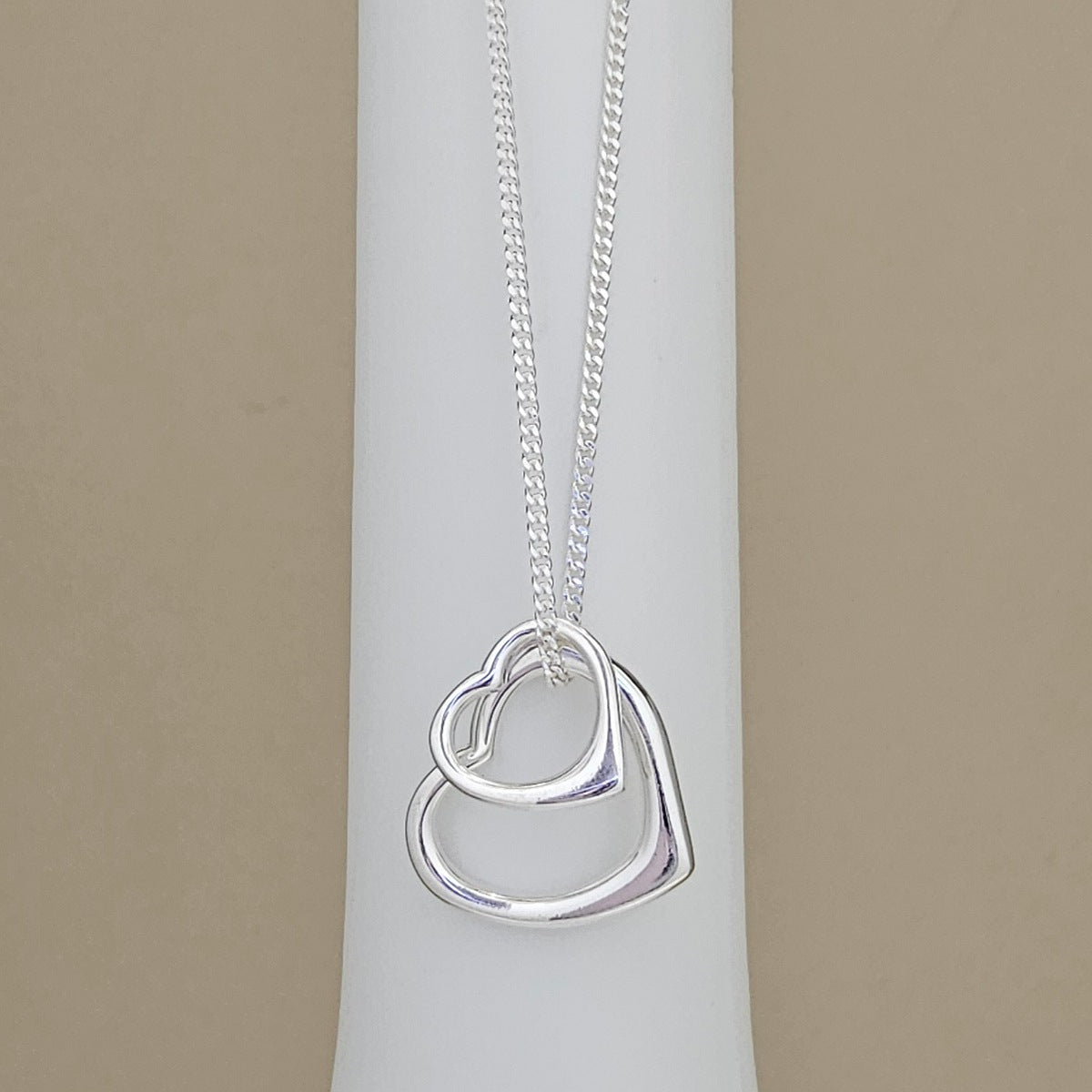 A silver necklace with a double interlocking heart pendant displayed on a white cylindrical jewelry stand against a beige background.