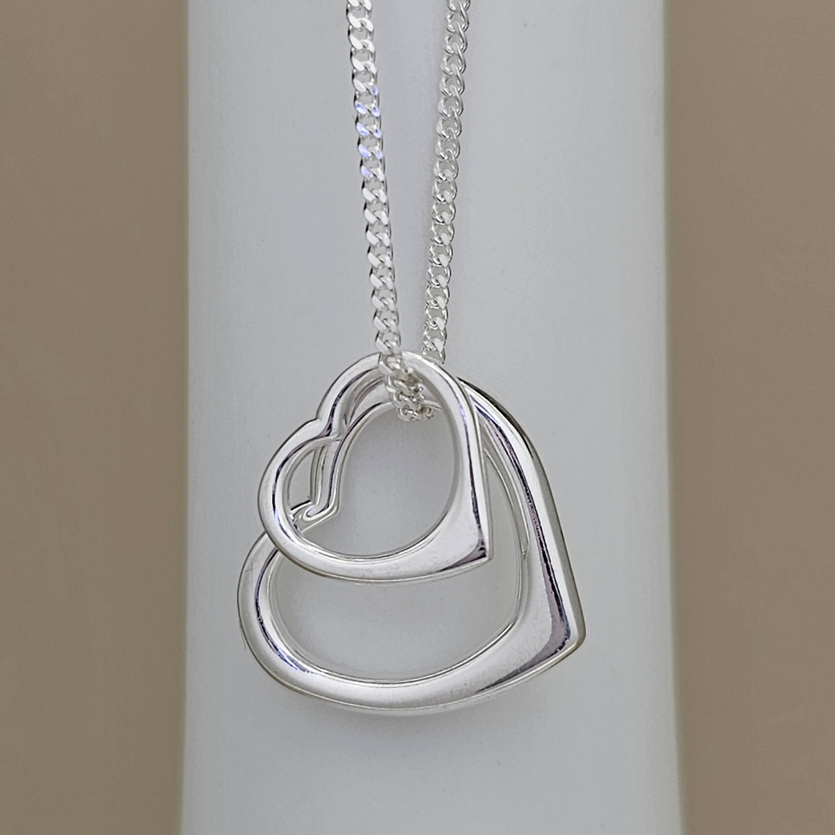 A silver double heart pendant necklace on a matching silver chain, displayed on a white cylindrical stand against a neutral beige background.