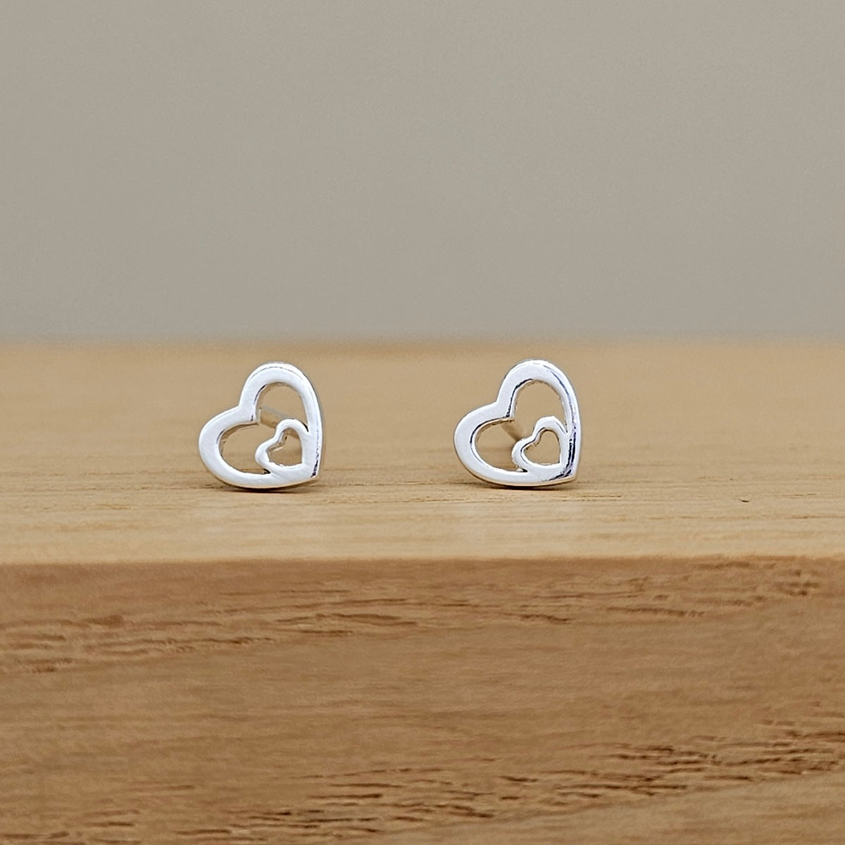 A pair of silver heart-shaped stud earrings with a smaller inner heart design, displayed on a light wooden surface against a neutral beige background.