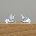 A pair of silver dove earrings with small gold accents, displayed on a wooden block against a neutral gray background.