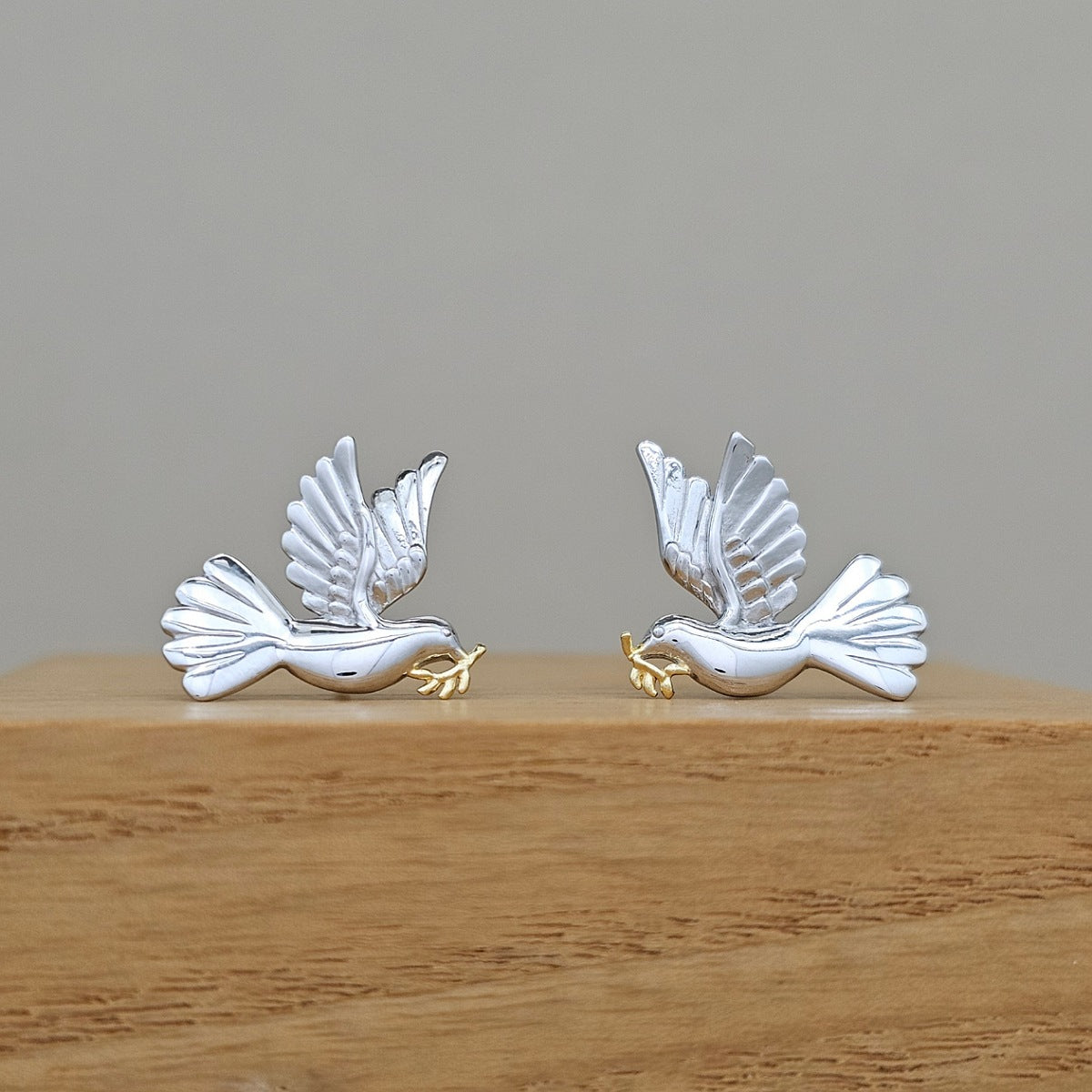 A pair of silver dove earrings with small gold accents, displayed on a wooden block against a neutral gray background.