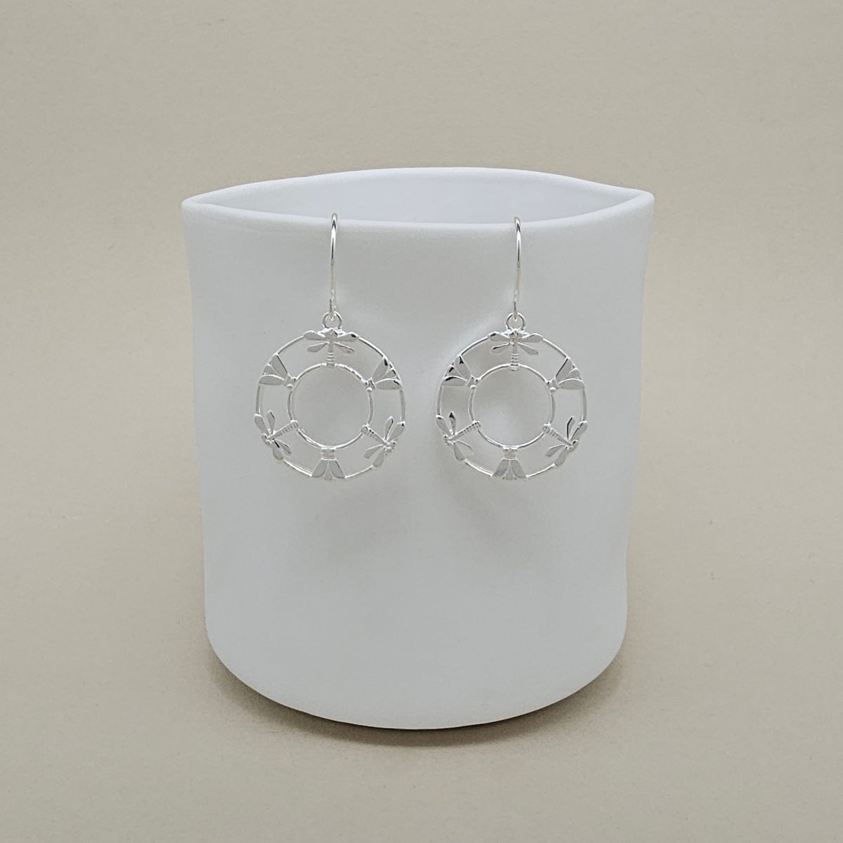 A pair of silver dangle earrings with circular openwork moth and dragonfly designs, displayed on a white ceramic cup against a warm beige background.