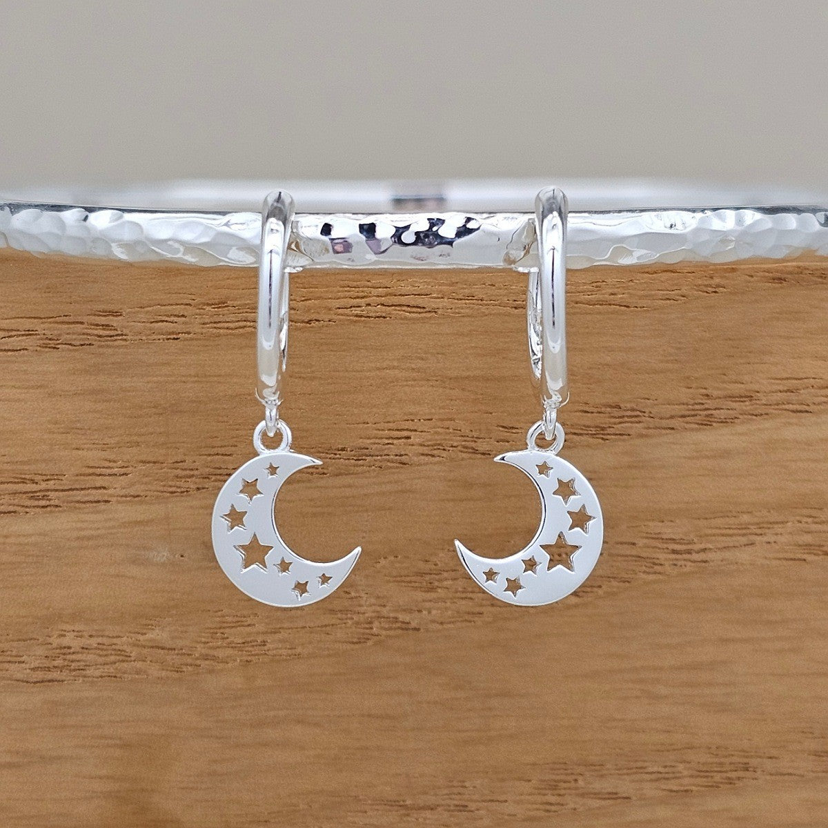 A pair of silver crescent moon and star dangle earrings hanging from silver hoop earrings, displayed on a textured silver bar over a wooden surface.