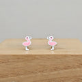 A pair of pink flamingo stud earrings with silver-toned metal outlines, displayed on a light wood block against a neutral gray background.