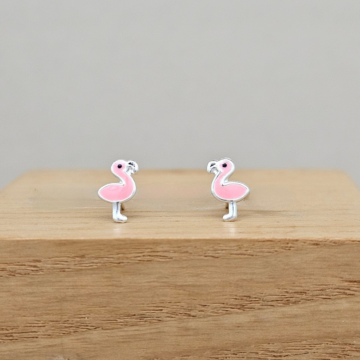 A pair of pink flamingo stud earrings with silver-toned metal outlines, displayed on a light wood block against a neutral gray background.
